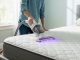 A person using a specialized mattress vacuum cleaner with UV-C light to sanitize mattress surface in a bright bedroom