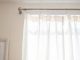 A metal curtain rod with brackets and rings holds a sheer white curtain against a beige wall.